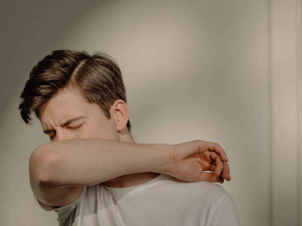 Mastering the First Impression: Your intriguing post title goes here A young man sneezes into his elbow, showcasing a hygiene practice, in an indoor setting.